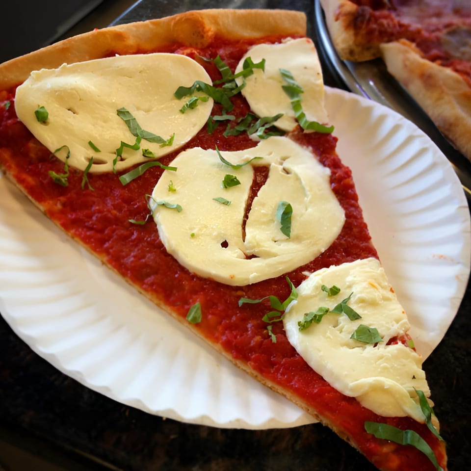 A slice of Nick's Pizza with fresh mozzarella, red sauce, and basil on a paper plate.