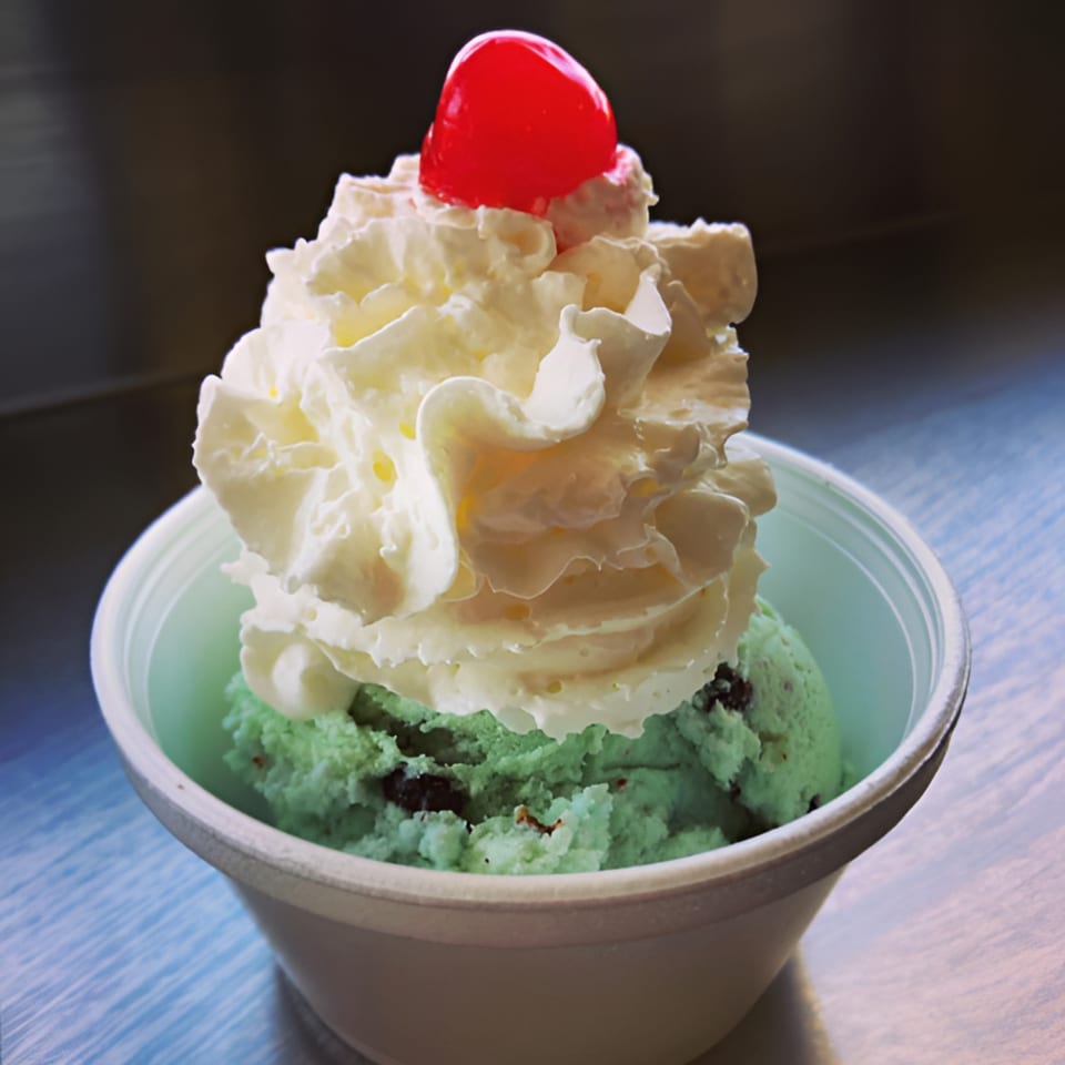 Mint chocolate chip ice cream with whipped cream and a cherry in a white cup.