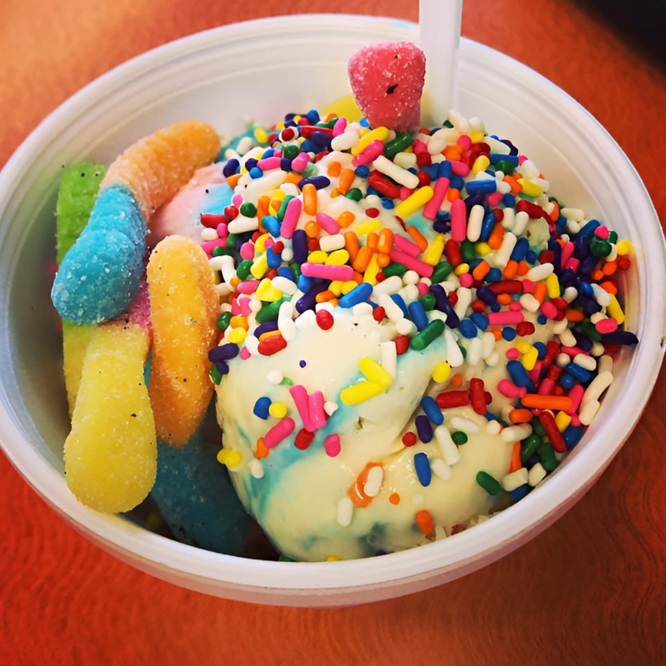 A white bowl of ice cream with colorful sprinkles, gummy worms, and a pink candy.